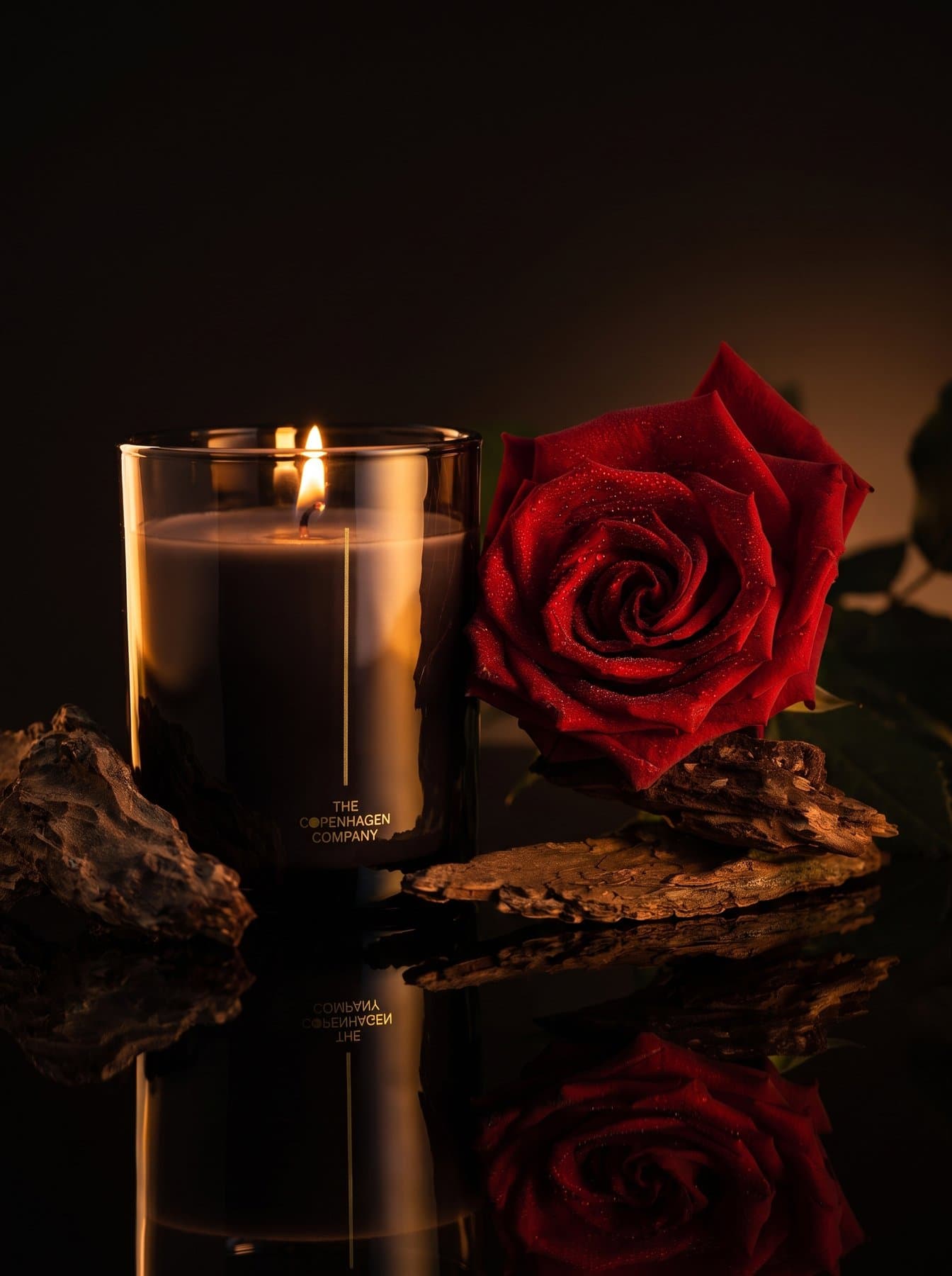 An Oud candle in dark glass beside a red rose